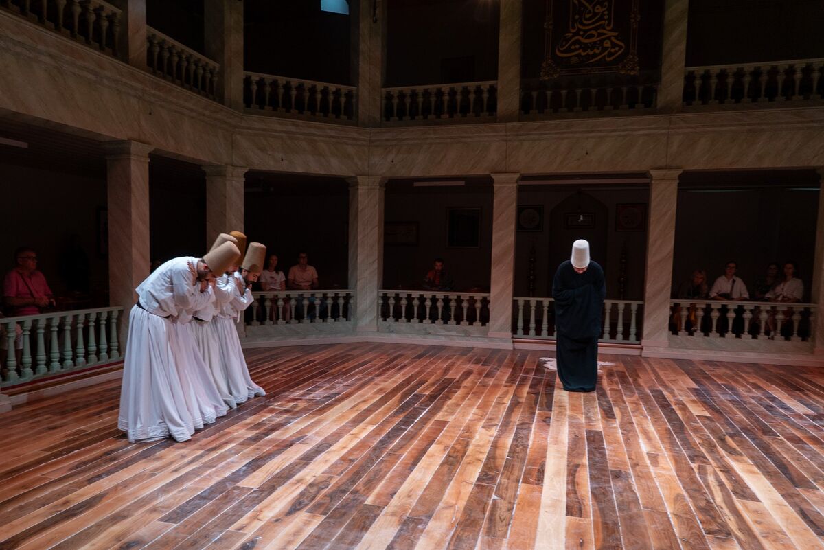 Four dervishes in white bowing in a line in the Kasımpaşa ceremony hall, a figure in a black cloak standing opposite them