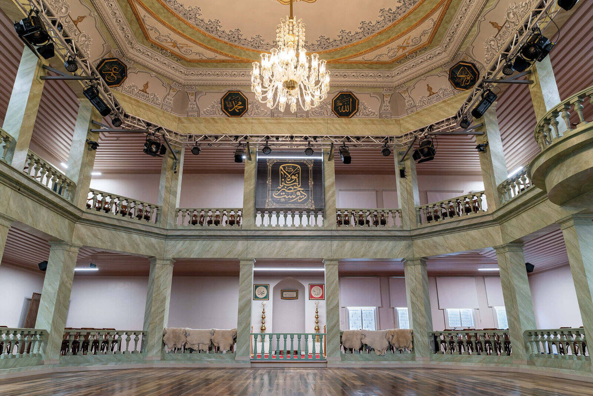 The empty octagonal ceremony hall of Kasımpaşa Mevlevihanesi with a wooden floor, a gallery on columns and a chandelier