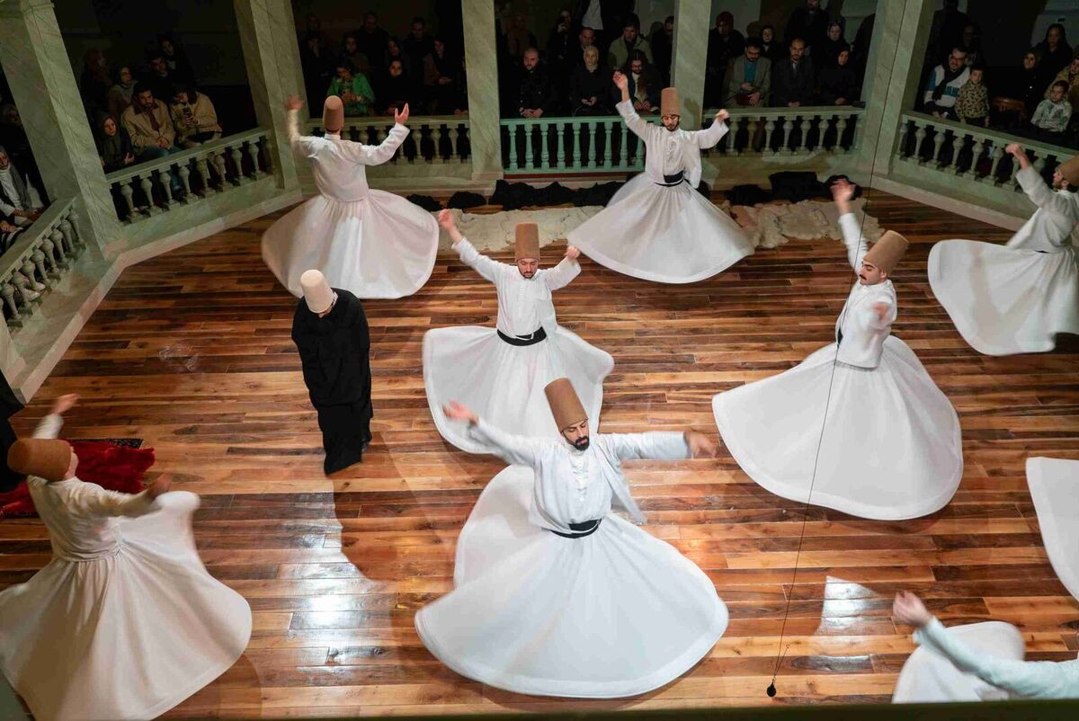 Whirling dervishes in white robes turning on the wooden floor of the ceremony hall at Kasımpaşa Mevlevihanesi, seen from the upper gallery